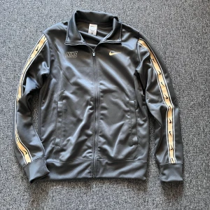 Grå träningsjacka från Nike - Säljer en snygg grå träningsjacka från Nike. Den har en dragkedja framtill och coola detaljer på ärmarna i guld och svart. Perfekt för träning eller som en casual vardagslook. Jackan är i bra skick och redo för nya äventyr!