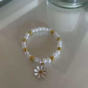 Pärlarmband med blomma - Säljer ett superfint armband med vita pärlor och guldfärgade detaljer. Armbandet har en söt blomma som hänge, perfekt för att ge en extra touch till din outfit. Passar både till vardag och fest!