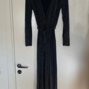 Svart glittrig långklänning - Säljer en elegant svart långklänning med glittriga detaljer. Klänningen har långa ärmar och ett knytband i midjan för en smickrande passform. Perfekt för festliga tillfällen!