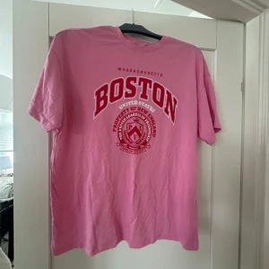 Rosa t-shirt från Divided med Boston-tryck - Säljer en superfin rosa t-shirt från Divided med ett coolt Boston-tryck i rött och vitt. Perfekt för en avslappnad stil eller när du vill ha något bekvämt men ändå snyggt. T-shirten är i bra skick och passar perfekt till jeans eller shorts. Skicka ett meddelande om du är intresserad!
