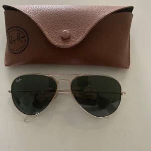 Ray-Ban pilotglasögon med fodral - Snygga Ray-Ban pilotglasögon i guld med mörka glas. Perfekt för en cool look året runt. Kommer med ett stilrent brunt fodral för säker förvaring. Glasögonen är i mycket bra skick och redo för nya äventyr!