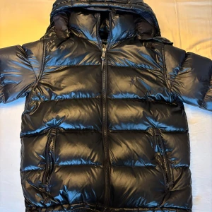 Svart dunjacka från Ralph Lauren (passar även L) - Säljer en snygg svart pufferjacka från Polo Ralph Lauren i storlek M. Jackan är perfekt för vintern med sitt quiltade mönster och långa ärmar. Den har en praktisk dragkedja framtill och är gjord i glansigt polyester-material. Huvan ger extra skydd mot kyla. Perfekt för kalla dagar! Den är köpt på NK förra vintern och kvitto finns kvar. Priset är inte skrivet i sten🙌🏼