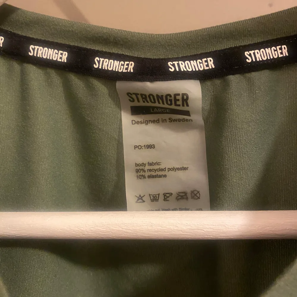 Säljer en grön kortärmad topp från Stronger i storlek L. Toppen är tillverkad av 90% återvunnen polyester och 10% elastan, vilket gör den både bekväm och stretchig. Perfekt för träning eller en avslappnad dag. Den har texten 'STRONGER' tryckt längs fållen.. Yläosat.