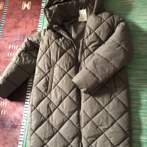 Olivgrön quiltad dunjacka - Säljer en olivgrön quiltad dunjacka i mycket bra skick. Den har en långärmad design med dragkedja och en mysig huva. Perfekt för kalla höst- och vinterdagar. Jackan är både stilren och funktionell, vilket gör den till ett måste i garderoben. Passar till både vardag och utflykter.