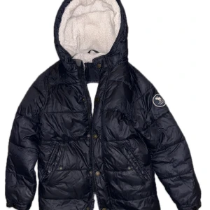 Svart pufferjacka från Abercrombie - Säljer en snygg svart pufferjacka från Abercrombie. Jackan har en mysig vit fleecefodring och en praktisk huva. Den är perfekt för kalla vinterdagar och har både dragkedja och knappar för extra skydd mot kylan.  Pris kan diskuteras 
