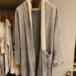 Grå morgonrock från Lounge Wear - Mysig grå morgonrock från Lounge Wear i storlek S. Den har en fluffig insida och långa ärmar, perfekt för att hålla sig varm och bekväm hemma. Den har dock en ficka som är av men som följer med ifall man skulle vilja sy fast den. 