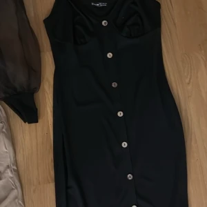 Svart klänning med knappar - Snygg svart klänning med knappar framtill. Den har en elegant och stilren design med en tight passform. Perfekt för festliga tillfällen eller en utekväll. Klänningen är ärmlös och har en klassisk look som aldrig går ur stil.