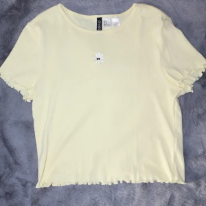 Gul t-shirt med broderad blomma. - Gul t-shirt med en liten broderad blomma på bröstet. T-shirten har korta ärmar och en lätt vågig kant vid ärmslut och nederkant. Aldrig använd.
