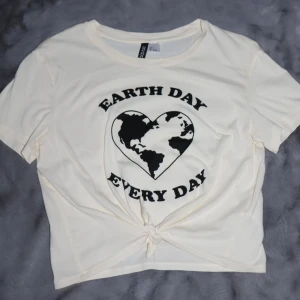 Vit Earth Day t-shirt - Vit t-shirt med texten 'Earth Day Every Day' och ett hjärta med jordglobsmotiv. Den har en knytning framtill. Den är i bra skick.