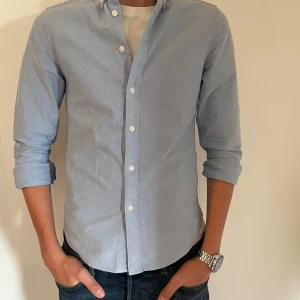 Ljusblå skjorta från East West - Säljer en stilren ljusblå skjorta från East West. Storlek S men passar på mig som är 180cm. Skjortan har en slim fit passform och är tillverkad i 100%bomull. Passar bra till vardag om man har en T-shirt under men är även bra om man ska klä upp sig