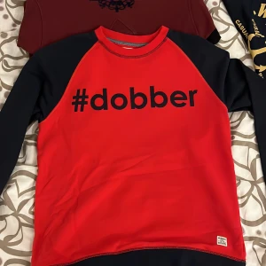 Röd och svart tröja från Dobber - Säljer en cool röd tröja från Dobber med svarta ärmar och hashtag-tryck på framsidan. Perfekt för en avslappnad stil. Tröjan har en normal passform och är gjord i mjukt material för extra komfort. Passar perfekt till jeans eller joggers! Passar M