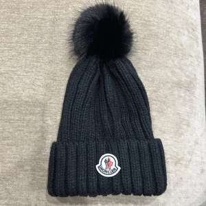Svart stickad mössa från Moncler - Säljer en helt ny snygg svart stickad mössa från Moncler med en fluffig äkta pälsboll på toppen. Den har en ribbad design och Moncler-loggan framtill. Perfekt för att hålla sig varm och stilren under vintern! Ej orginal. 