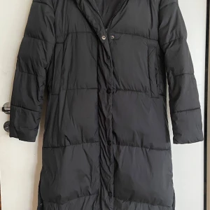 Svart lång dunjacka från Everest - Säljer en svart lång dunjacka från Everest i storlek 34. Perfekt för kalla vinterdagar med sin varma och bekväma design. Jackan har långa ärmar och stängs med knappar. Den är i mycket bra skick och redo för nya äventyr!