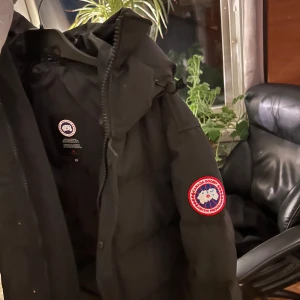Svart dunjacka från Canada Goose - Säljer en svart dunjacka från Canada Goose i mycket bra skick. Den är perfekt för kalla vinterdagar med sin varma och skyddande design. Jackan har en klassisk look med en broderad logga på ärmen och dragkedja framtill. Perfekt för dig som vill hålla stilen i vinterkylan! 