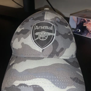 Camouflage keps med Arsenal-logga - Snygg keps i camouflage-mönster med Arsenal-logga framtill. Perfekt för fotbollsfans som vill visa sitt stöd med stil. Kepsen har en klassisk passform och justerbar rem baktill för bästa passning.