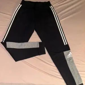 Säljer ett par snygga mjukisbyxor från Adidas i svart och grått. De har de klassiska tre vita ränderna längs sidorna och en grå sektion med Adidas-loggan på benet. Perfekta för träning eller chill hemma. Superbekväma och i bra skick!