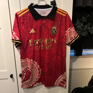 Röd fotbollströja från Adidas - Säljer en snygg röd fotbollströja från Adidas med Real Madrid-logga och drakmönster. Tröjan har korta ärmar och en svart krage. Perfekt för fans som vill sticka ut på matchen! 🐉