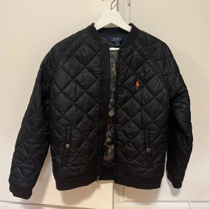 bomberjacka från Ralph Lauren - Säljer en snygg svart quiltad bomberjacka från Polo Ralph Lauren. Perfekt för höst och vår med sin stilrena design och bekväma passform. Passar både till vardags och fest!