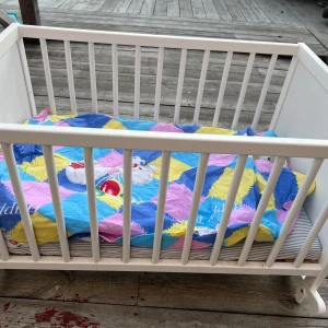 Vit spjälsäng med färgglad filt - Säljer en söt vit spjälsäng i trä med hjul för enkel förflyttning. Den har en färgglad filt i blått, gult, rosa och lila med ett lekfullt mönster. Perfekt för barnrummet! 🍼