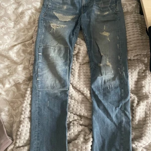 G-Star Jeans - Säljer ett par coola slitna jeans i blått. De har en straight passform och är i bra skick med snygga slitningar på framsidan och baksidan. Perfekta för en avslappnad stil!