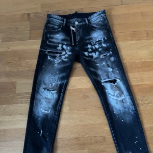 Svarta jeans från Dsquared2 - Säljer ett par svarta Dsquared2 jeans i storlek 40. De har en cool distressed look med slitningar och färgstänk. Jeansen har en dragkedja och knapp framtill. Perfekta för en edgy stil!