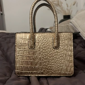 Guldig handväska från H&M - Snygg guldig handväska från H&M med krokodilpräglat mönster. Perfekt för att ge din outfit en lyxig touch. Väskan har en praktisk dragkedja och två handtag för enkel bärning. Den är i mycket bra skick och passar perfekt för både vardag och fest.