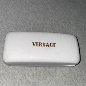 Solglasögon från Versace - Snygga solglasögon från Versace med en elegant design. De har en svart och guld färgkombination med en distinkt logotyp på framsidan. Glasögonen har en rektangulär form och kommer i ett stilrent vitt fodral med guldfärgad text.
