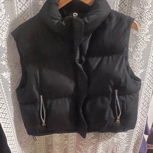 Svart dunväst med dragkedjor - Snygg svart dunväst med dragkedjor på fickorna. Perfekt för kyligare dagar och ger en cool look. Västen har en hög krage och en stilren design. Den är från newyorker