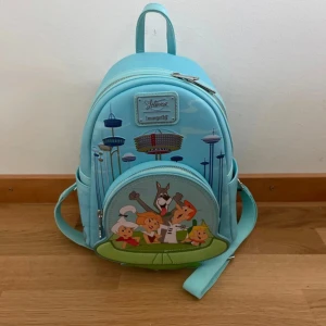 Blå ryggsäck med Jetsons-motiv från Loungefly - Säljer en cool blå ryggsäck från Loungefly med Jetsons-motiv. Väskan har ett huvudfack och en framficka, båda med dragkedja. Perfekt för dig som älskar retro och tecknade serier! 🎒