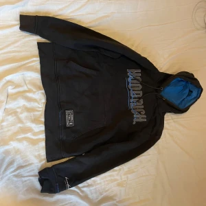 Svart hoodie från Hoodrich - Säljer en svart hoodie från Hoodrich med blått foder i huvan och tryck på både fram- och baksidan. Tröjan har en klassisk känguruficka och ribbade muddar vid ärmslut. Perfekt för en avslappnad stil.