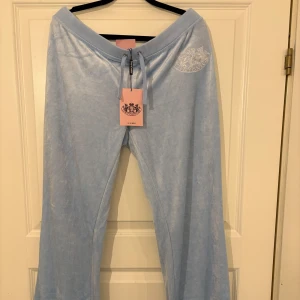 Ljusblå byxor från Juicy Couture - Snygga ljusblå byxor från Juicy Couture med broderad detalj på framsidan. Byxorna har en bekväm passform med snörning i midjan och fickor med knappar baktill. Perfekta för en avslappnad stil. Aldrig använda, prislappen är fortfarande kvar på byxorna🥰