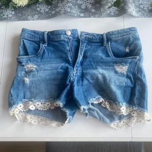 jeansshorts med spetsdetaljer - Snygga blå jeansshorts med slitna detaljer och vacker spetskant längst ner. Perfekta för en avslappnad sommarlook. De har en klassisk femficksdesign och knappgylf.