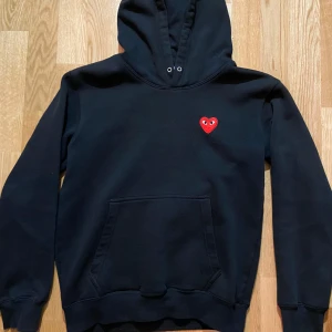 Svart cdg play hoodie  - Säljer en stilren svart hoodie med en röd hjärtdetalj på bröstet. Tröjan har en klassisk känguruficka och en bekväm huva med dragsko. Perfekt för en avslappnad look.
