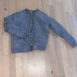Blå bomberjacka - Säljer en stilren blå bomberjacka med dragkedja och ribbade muddar. Jackan har långa ärmar och en klassisk bomberdesign med fickor och detaljerade sömmar. Perfekt för en cool och avslappnad look. Från 11-12 åringar! Den har tyvärr en liten Skråma dock men man kan säkert få bort den enkelt.