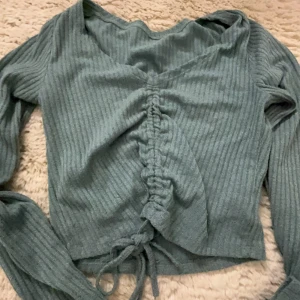 Grön ribbad tröja med knyt - Säljer en snygg grön ribbad tröja med v-ringning och knyt framtill. Tröjan har långa ärmar och en trendig design som passar perfekt för både vardag och speciella tillfällen.