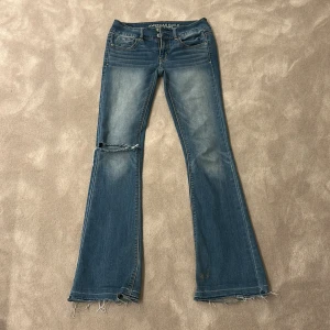 Blå bootcut jeans från American Eagle - Snygga blå bootcut jeans från American Eagle med slitna detaljer och låg midja. Perfekta för en avslappnad stil. De har en klassisk femficksdesign och en bekväm passform.