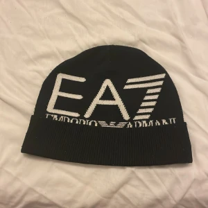 Svart mössa från Emporio Armani - Snygg svart mössa från Emporio Armani med stor vit logga 'EA7' framtill. Perfekt för att hålla värmen med stil. Mössan är ribbad nedtill för extra passform.