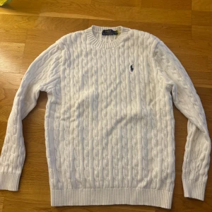 Vit stickad tröja från Ralph Lauren - Säljer en klassisk vit stickad tröja från Ralph Lauren med kabelstickat mönster och den ikoniska loggan broderad i blått på bröstet. Tröjan har långa ärmar och ribbstickade muddar vid ärmslut och nederkant. Perfekt för kyliga dagar!