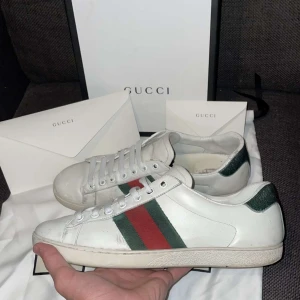 Vita sneakers från Gucci - Säljer ett par vita sneakers från Gucci med de ikoniska gröna och röda ränderna på sidan. Skorna har snörning och en klassisk design som passar perfekt för en stilren look. Allt OG ingår!!