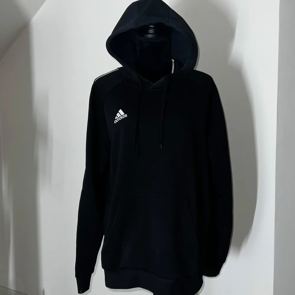 Säljer en stilren svart hoodie från Adidas med deras klassiska logga på bröstet. Tröjan har en bekväm huva med dragsko och en stor magficka framtill. Perfekt för en avslappnad stil.. Hupparit & Collegepaidat.