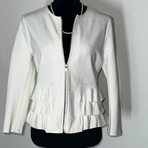 Vit jacka med volangdetaljer - Elegant vit jacka med dragkedja framtill och volangdetaljer på fickorna. Jackan har en stilren design med långa ärmar. Perfekt för en chic och sofistikerad look.