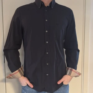 Svart skjorta från Burberry Brit - Snygg svart skjorta från Burberry Brit med klassiska rutiga detaljer på insidan av manschetterna. Skjortan har långa ärmar och knappar framtill. Perfekt för en stilren look. Storlek L men passar bättre till M