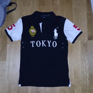 Chief Keef Polo - Snygg svart pikétröja från Ralph Lauren med vita ärmar och röda detaljer. Tröjan har en broderad logga och texten 'TOKYO' på framsidan samt en stor siffra '5' på baksidan. Perfekt för en stilren look. EXTRAPRIS I 1 DAG