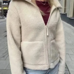 Beige teddyjacka med dragkedja - Mysig beige teddyjacka med dragkedja framtill. Perfekt för kyliga/vår dagar med sin fluffiga och varma design. Jackan har en enkel och stilren look som passar till det mesta.