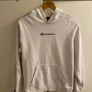 Vit hoodie från Champion - Säljer en vit hoodie från Champion med svart logga på bröstet och längs ärmarna. Tröjan har en klassisk känguruficka och en bekväm huva. Perfekt för en avslappnad stil.