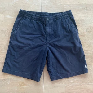 Marinblå shorts från Polo Ralph Lauren - Snygga marinblå shorts från Polo Ralph Lauren med elastisk midja och knappstängning. De har två sidofickor och två bakfickor med knappar. Broderad logga i vitt på benet ger en stilren touch.