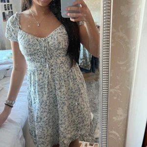 Blommig klänning med knyt - Blommig klänning i ljusa färger med korta ärmar och knyt framtill. Den har en smickrande passform med elastisk midja som ger en bekväm känsla