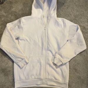 Vit hoodie från FSBN - Säljer en vit hoodie från FSBN i storlek L. Den har en dragkedja framtill och en mysig huva. Perfekt för en avslappnad stil. Slim fit passform för en modern look.