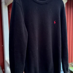 Svart tröja från Ralph Lauren - Säljer en stilren svart tröja från Ralph Lauren med det klassiska röda logotypbroderiet på bröstet. Tröjan har en rund halsringning och är tillverkad i ett texturerat material som ger en lyxig känsla. Perfekt för en elegant och avslappnad look.             Skriv vid eventuella frågor! Pris kan Alltid diskuteras!