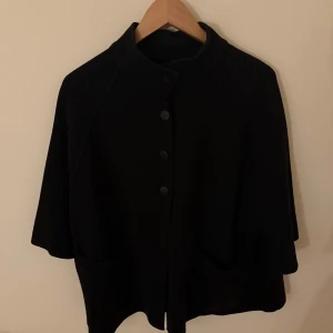 Svart kappa med knappar - Snygg svart kappa med knappar framtill. Den har en stilren design och är perfekt för kyligare dagar. Passar bra över en tröja för extra värme.